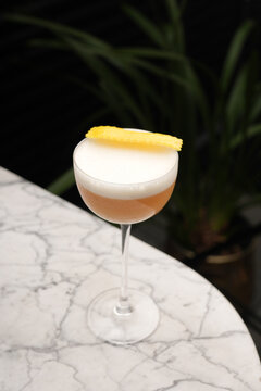 Classic sour cocktail with thick white foam and a yellow lemon twist garnish, served in a coupe glass on a marble table against a dark background. Fine dining bar drink