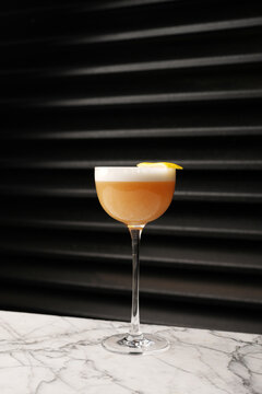 Classic sour cocktail with thick white foam and a yellow lemon twist garnish, served in a coupe glass on a marble table against a dark background. Fine dining bar drink