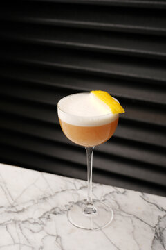 Classic sour cocktail with thick white foam and a yellow lemon twist garnish, served in a coupe glass on a marble table against a dark background. Fine dining bar drink