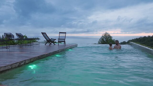 A devoted father and his young son are enjoying precious quality time in their stunning swimming pool, which is beautifully surrounded by scenic views and the calming tranquility of nature