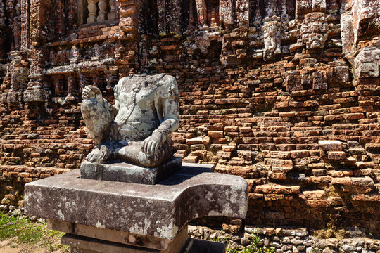 Ancient headless stone statue seated on a pedestal outside the brick walls of Group D at My Son Sanctuary