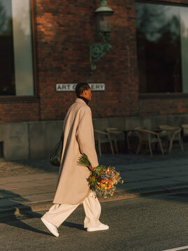 Elegant Woman in Beige Trench Coat Walking on European City Street Holding Delicate Wildflowers in Golden Autumn Sunlight