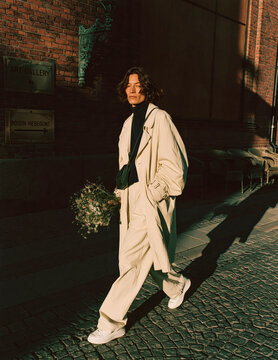 Elegant Woman in Beige Trench Coat Walking on European City Street Holding Delicate Wildflowers in Golden Autumn Sunlight