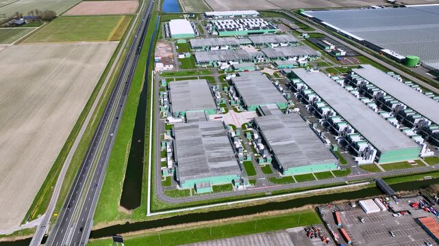 Aerial view of a massive industrial data center facility with cooling units and flat agricultural fields under a clear sky in Middenmeer, North Holland, Netherlands.