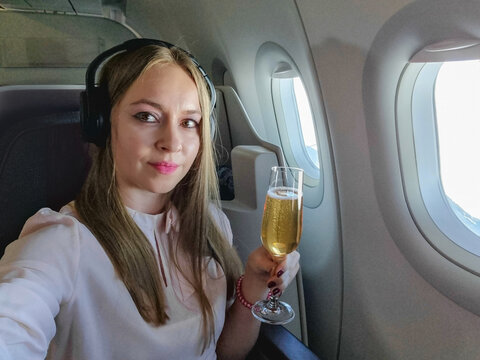 Woman passenger enjoying a glass of champagne while traveling in a premium airline cabin. Relaxing at a window seat with headphones on during a long-haul flight. Concept of luxury travel, business cla