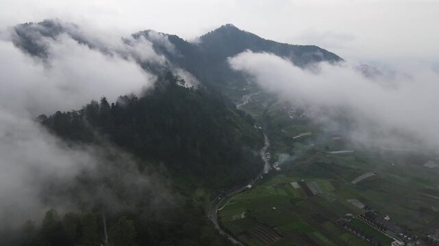 Aerial view of lush green mountains and terraced fields shrouded in thick white clouds and mist over a winding road, Tawangmangu, Central Java, Indonesia.