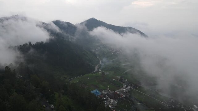 Aerial view of thick white clouds rolling over lush green mountains and a small village settlement in the valley of Tawangmangu, Central Java, Indonesia.