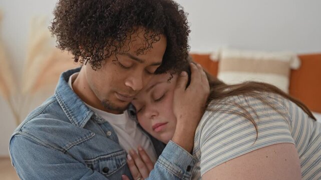 Man cradles woman's face and holds her head while she leans on his chest on a bed in a studio; comfort healing.