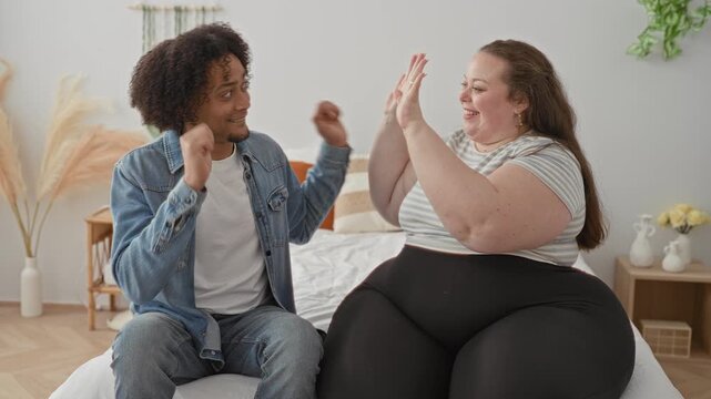Man and plus size woman clasp hands and high five on bed, smiling in a cozy bedroom inside a building; affection connection.