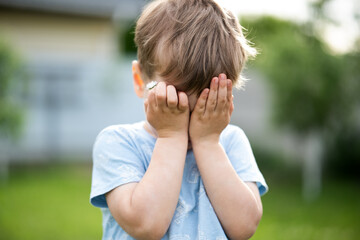 Fototapeta premium Shy little boy covering his face with hands while holding a daisy flower in a sunny garden. Childhood emotions, innocence, playfulness, outdoor lifestyle and tender moments.
