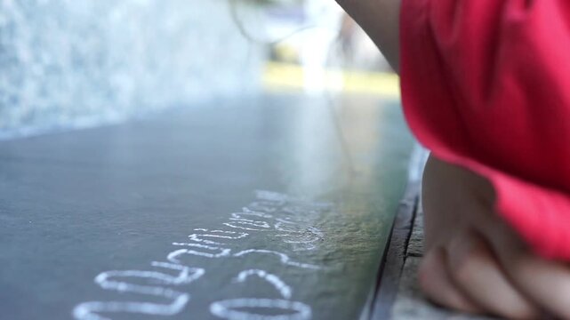 Close-up shot of a left-handed child wearing a red shirt drawing wavy lines and writing with a piece of white chalk on dark outdoor floor tiles during a bright day.