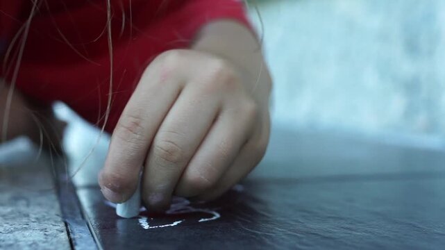 ​Wide shot of a young boy sitting on the pavement, deeply focused on drawing and writing with colorful chalks during a bright summer afternoon.