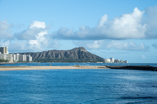 Diamond Head, a famous volcanic tuff cone, stands prominently behind the Waikiki coastline with its modern buildings and a sandy beach meeting the clear blue Pacific Ocean