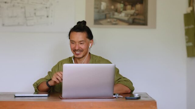 Creative professional with top knot hairstyle wearing olive green linen shirt participates in video conference call using laptop and earphones at wooden desk in minimalist home office space.