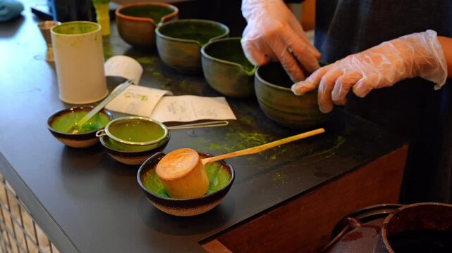 Matcha tea preparation with ceramic bowls and bamboo tools. Gloved hands arranging dark stoneware on countertop with green powder and sieve.
