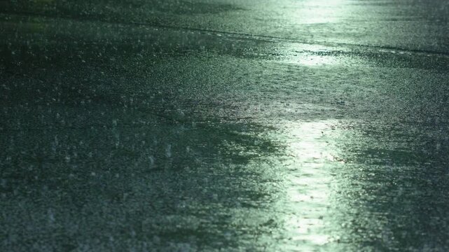 Heavy rain pours down onto dark wet asphalt road surface at night. Raindrops splash and create ripples on the flooded pavement while streetlight reflections shimmer across the glistening wet ground.