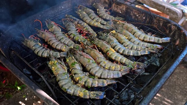 Large tiger prawns cooking on a charcoal barbecue grill. Overhead perspective of seasoned seafood pieces smoking over hot coals with green herbs and garlic marinade.