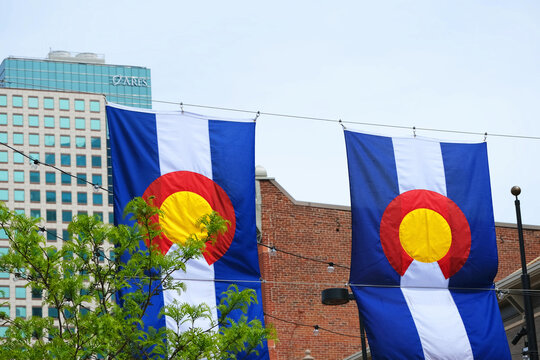 Two Colorado state flags hung across a downtown street between brick and glass buildings.