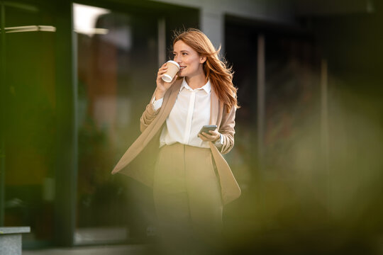 Businesswoman drinking coffee and using smartphone while walking near office building