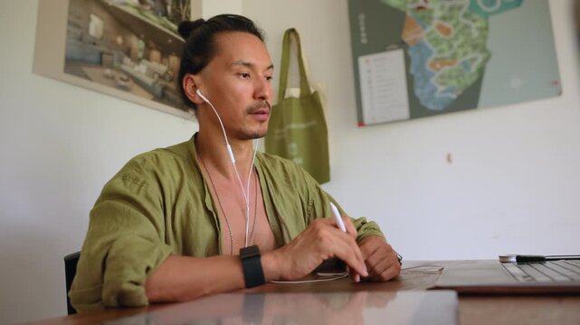 Business video call session with focused man wearing earphones while working on laptop at wooden desk. He holds stylus pen and wears casual green linen shirt in modern home office with map on wall.