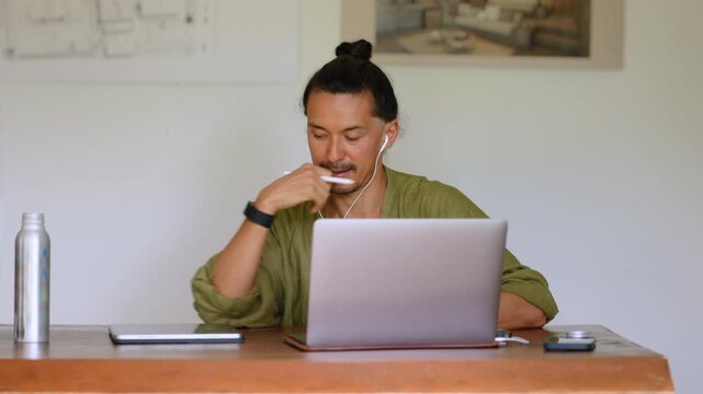Creative professional man with hair bun wearing green linen shirt participates in video conference call on laptop while taking notes with stylus on tablet at wooden desk in minimalist home office.
