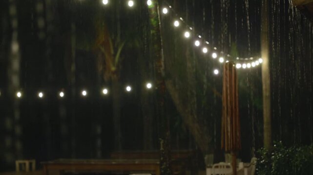 Heavy rain pours down on tropical outdoor patio decorated with glowing string lights. Raindrops fall through warm illumination as palm trees and wooden furniture create cozy atmosphere in darkness.