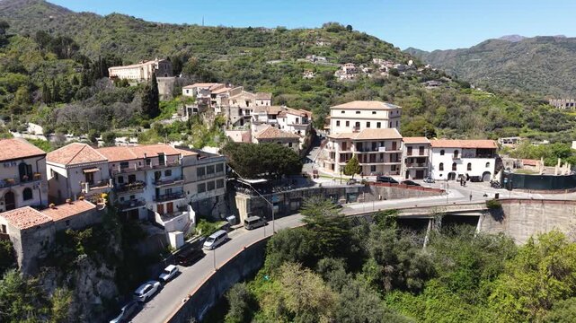Aerial view of the historic village of Savoca with stone houses and terracotta roofs nestled on a lush green hillside under a clear blue sky in Savoca, Sicily, Italy.