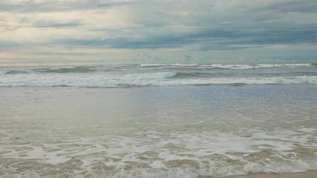Gentle ocean waves roll onto a sandy beach under a dramatic overcast sky. White foam spreads across wet sand as multiple wave lines break in the distance. Calm seascape scene.