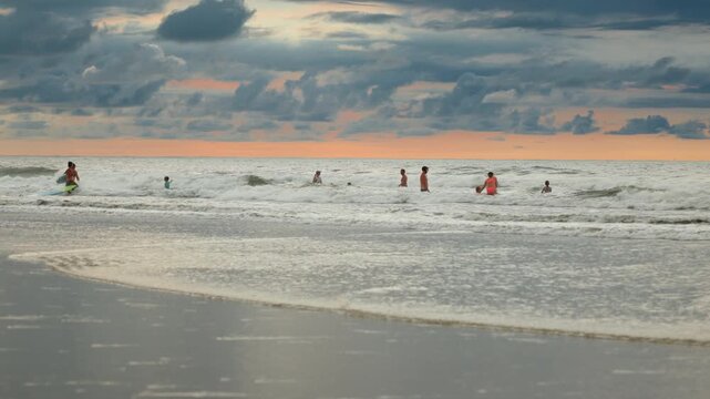 Group of beachgoers enjoying ocean waves during golden hour sunset. Dramatic clouds fill the sky with pink and orange hues as swimmers play in the foamy surf on sandy shoreline.