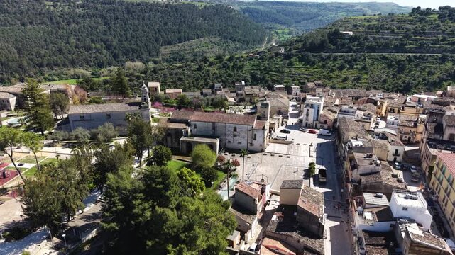 Aerial view of the historic stone buildings, central square, and church bell tower surrounded by lush green hills in Ragusa, Sicily, Italy.