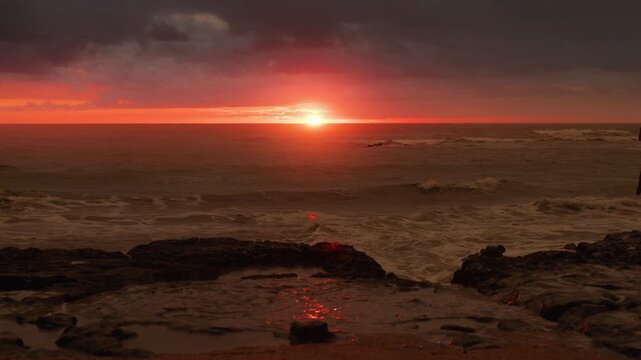 Breathtaking sunset over ocean with golden sun on horizon casting orange and red light across dark stormy clouds. Waves crash against rocky shore with tide pools reflecting warm colors.