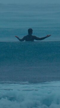 Surfer sitting under stormy rain waiting for wave, dark teal ocean and distant swell, silhouette with arms outstretched, heavy drizzle and foam churn, moody dusk atmosphere, vertical video
