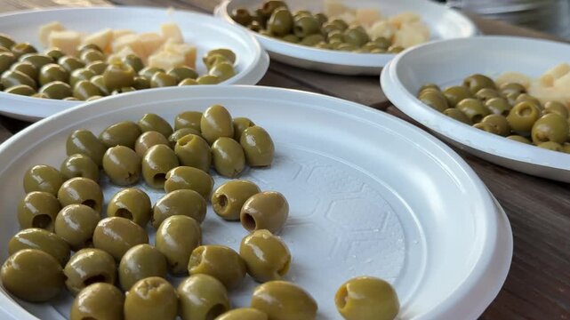 Festive outdoor picnic food with pitted olives and parmesan cheese cubes arranged on plastic plates for barbecue gathering in nature.