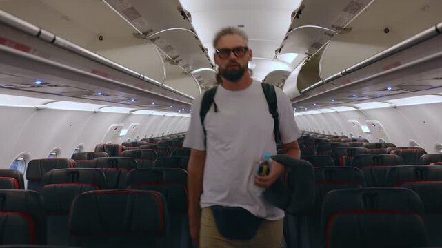 Bearded man wearing glasses and white t-shirt walks through empty commercial airplane cabin aisle carrying backpack and hat. Rows of dark seats with red trim visible on both sides.