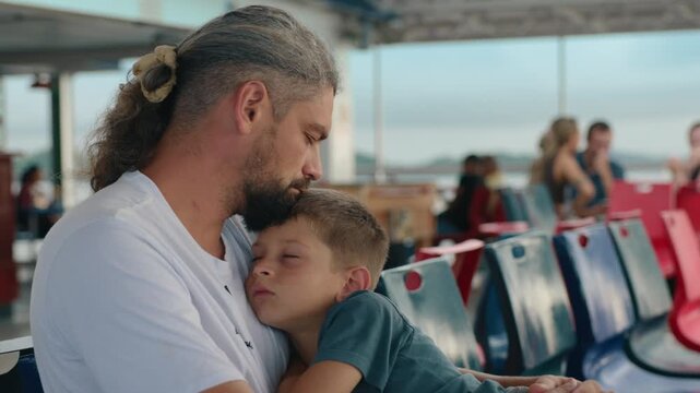 Father and son dozing off on a ship as the boy sleeps peacefully in his dads arms on colorful ferry seats. Passengers in background enjoy the sea voyage during warm summer travel.