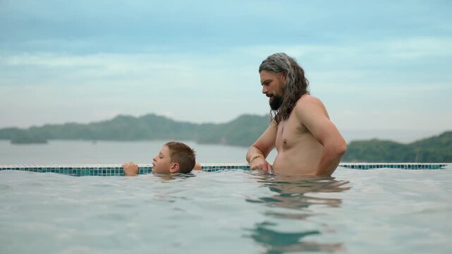 Father and son share quality time in stunning infinity pool with turquoise blue tiles, calm water reflecting cloudy sky, lush green tropical mountains visible in misty distance on horizon.
