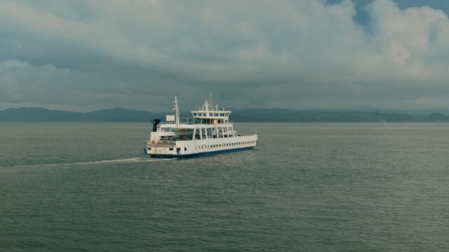 Aerial view of white passenger car ferry navigating across calm green ocean waters. Dramatic cloudy sky above with distant mountainous coastline visible on the horizon. Maritime transportation scene.