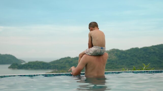 Father holds young son on shoulders while standing in infinity pool overlooking lush green mountains and ocean bay. Peaceful family vacation moment at tropical resort with scenic landscape view.