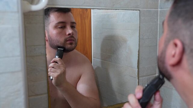 A young man trimming his beard with a trimmer in the bathroom in front of the mirror