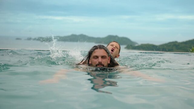 Bearded father swims with young son on his back in turquoise infinity pool. Boy splashes water while dad carries him with tropical mountains and cloudy sky in background.