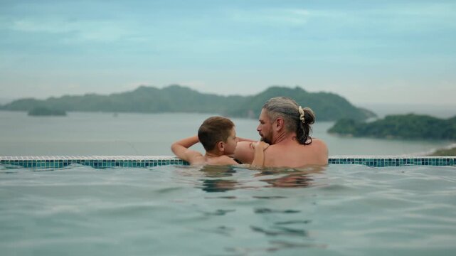 Father and young son share a bonding moment in an infinity pool overlooking tropical islands and mountains. The man with gray hair holds his child while gazing at the scenic ocean vista.