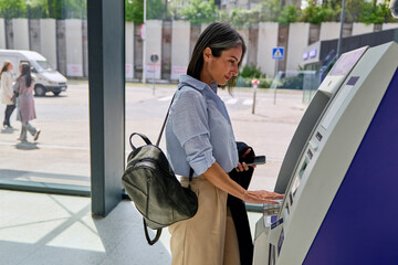Obraz premium Woman using a self service banking machine on a city street, managing finances with smartphone and backpack, efficient modern digital transaction in everyday urban life