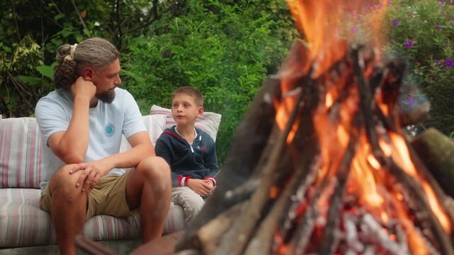 Father and son enjoying quality time by campfire in lush garden setting. Bearded dad listens attentively as boy talks animatedly while flames dance in foreground creating warm cozy atmosphere.