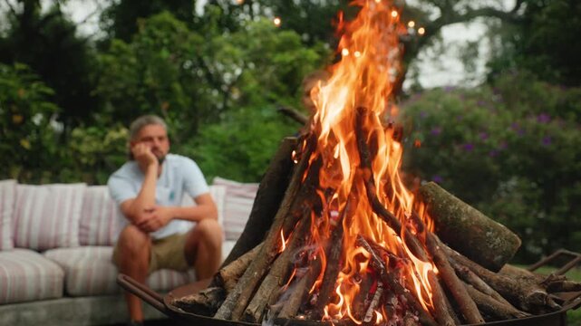 Father and son enjoy quality time around a blazing backyard fire pit at dusk. Boy adds sticks to the flames while dad watches from a striped cushioned outdoor sofa surrounded by lush green trees.