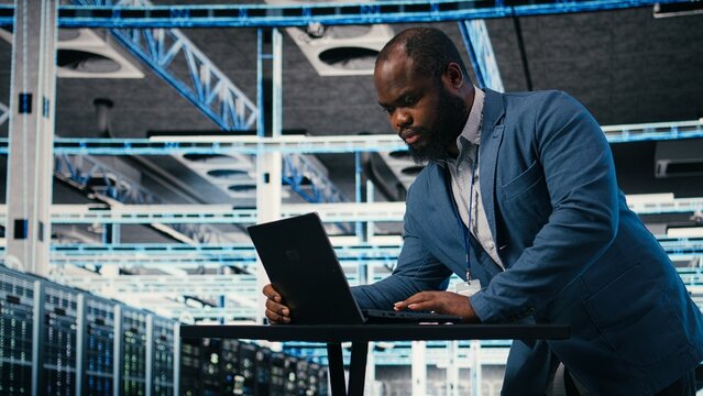 Programmer at server farm workplace desk using laptop, doing network management, Data center IT expert overseeing IT infrastructure supercomputers with software, typing on notebook keyboard, camera A.
