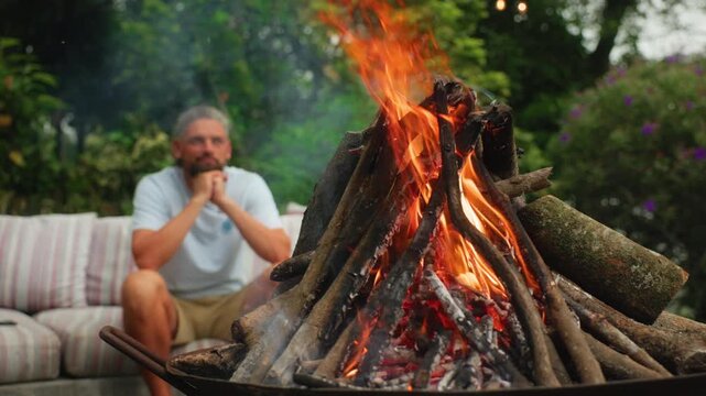Middle-aged man with gray hair sits pensively on striped outdoor sofa watching flames dance in metal fire pit. Burning logs and glowing embers in foreground with green garden backdrop.
