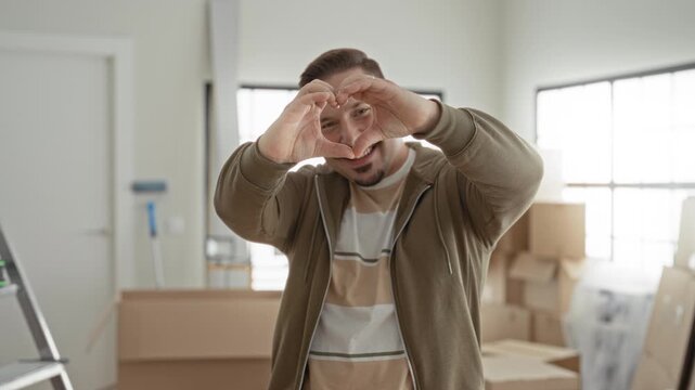 Young hispanic man forms heart with hands amid stacked moving boxes in an apartment building near a ladder and bright window light; home love.