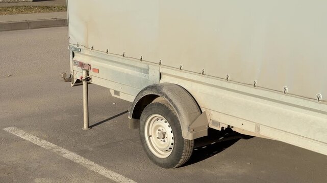 Covered passenger trailer parked in lot during spring sunlight gray tarpaulin fastened over cargo bay single axle wheel, hitch