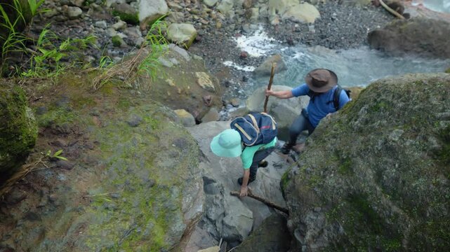 Father and son hiking through wild nature as they navigate moss-covered boulders beside a rushing mountain stream. Adult guides child across rocky terrain with walking stick in lush wilderness.