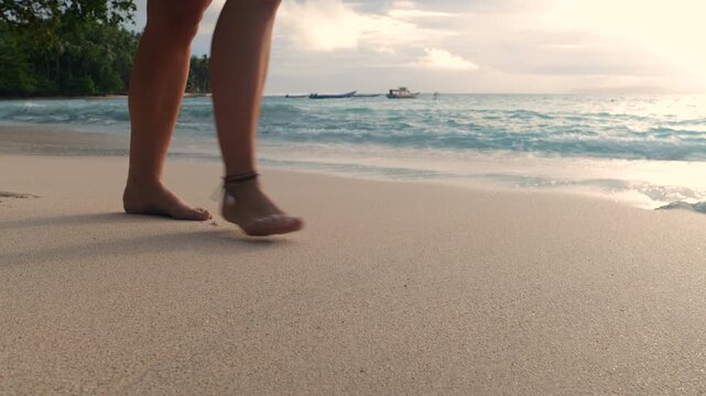 SLOW MOTION, LENS FLARE, CLOSE UP: Female bare feet walking across soft sand as gentle waves roll in under sunset light. Shoreline with palms and distant boats add a serene mood of Mentawai Islands.